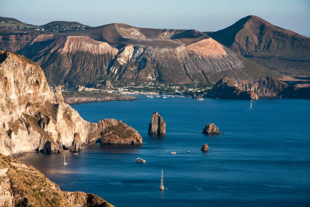 Image of Volcano Island (Baia di Levante) in Italy where the cyanobacterial strains were isolated. Image contains rock formations and a body of water in the foreground with more rock formations in the background. 